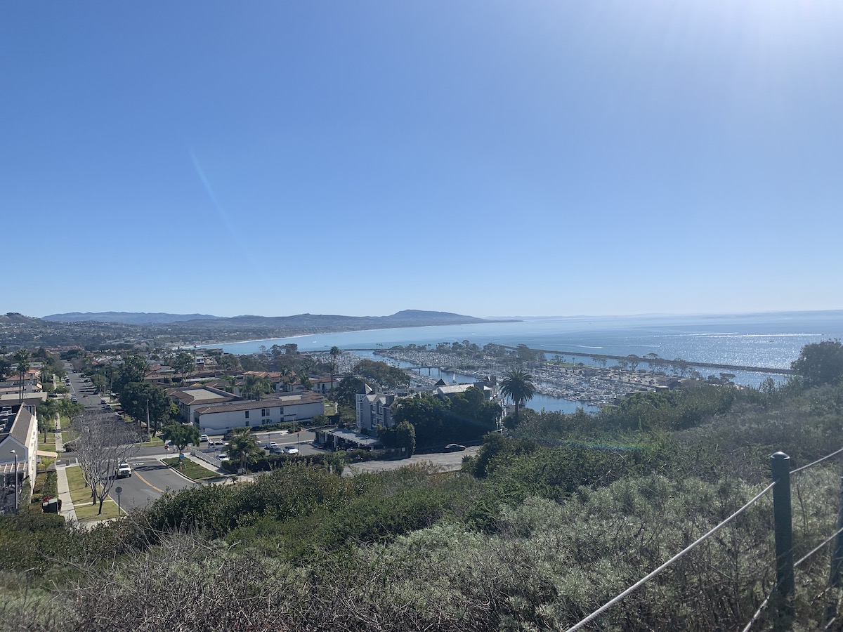 A landscape photo with the Pacific ocean on the right, a harbor in the middle and houses on the left. Mountains and more coast line are in the background.