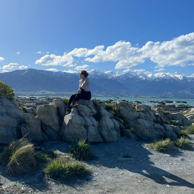 A white woman, Mac, sits on a large boulder with snow capped mountains and a bay in the distance