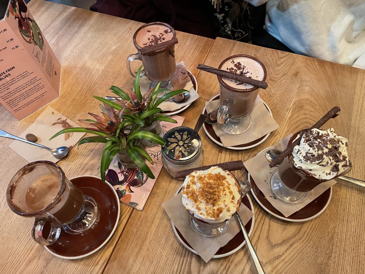 A wooden table with 5 decadent hot chocolates in glass cups with whipped cream or sprinkles on top from above