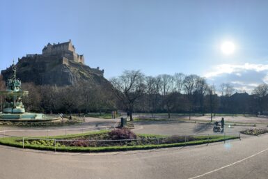 A panoramic view of Edinburgh Castle from Princes Garden, including a fountain and church, with the sun shining.