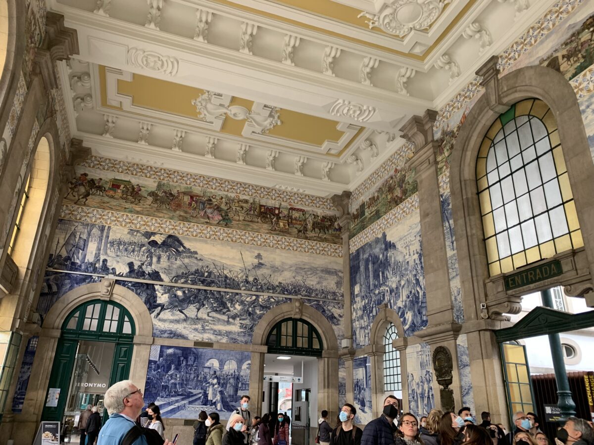 The main foyer of a train station with blue tiles on the walls forming art depicting the history of Porto with gold and white crown moulding on the ceiling