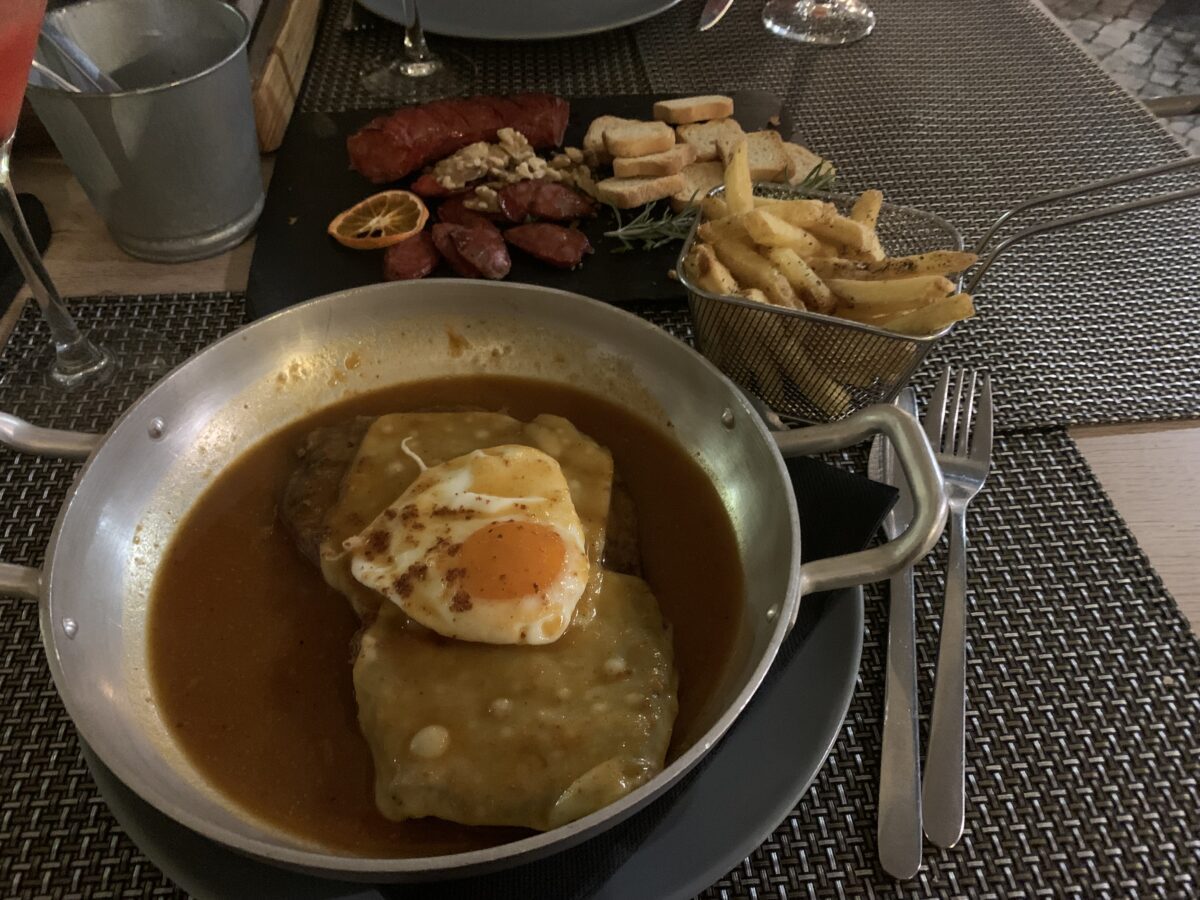 A metal bowl with gravy, two slices of cheese, and a fried egg on top of a steak, with fries and olives on the table behind