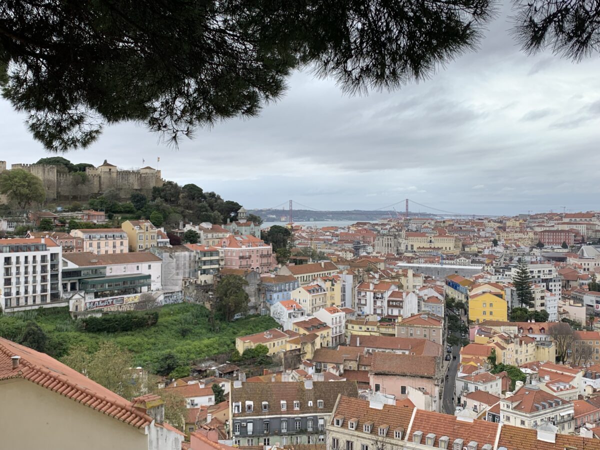 A panoramic view of Lisbon with buildings with red tiled roofs from end to end and the river with the bridge in the distance
