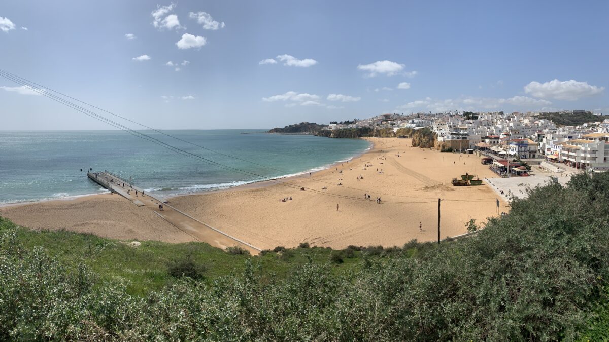A panoramic of a beach with a large beige sand beach, green bushes in the fore ground, and mostly white buildings stretching into the distance