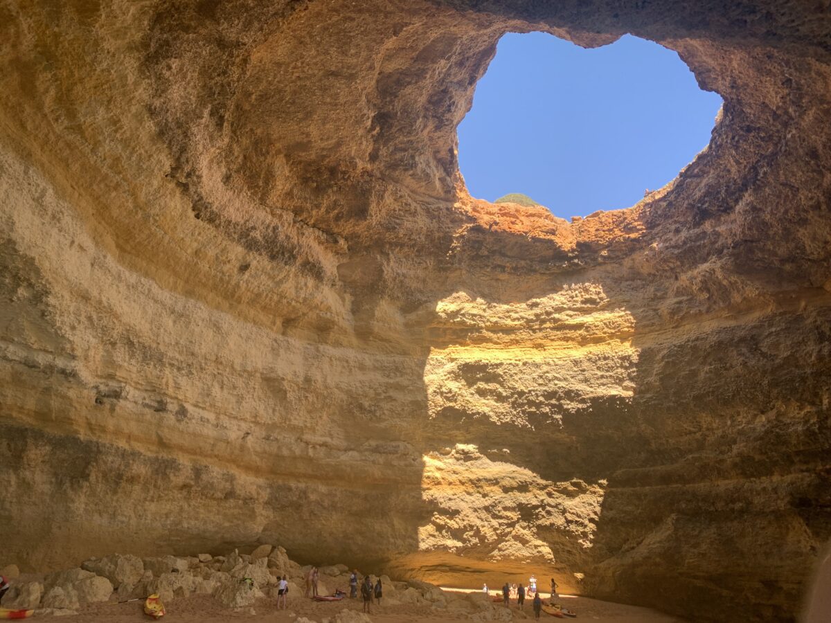 A large cave with golden earth walls and one large hole in the ceiling where a beam of sunlight comes through