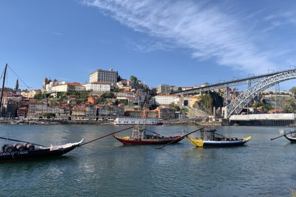 ships used to carry port are docked in the river with the hills of Porto city rising behind them and a large metal bridge reaching across the river on the right