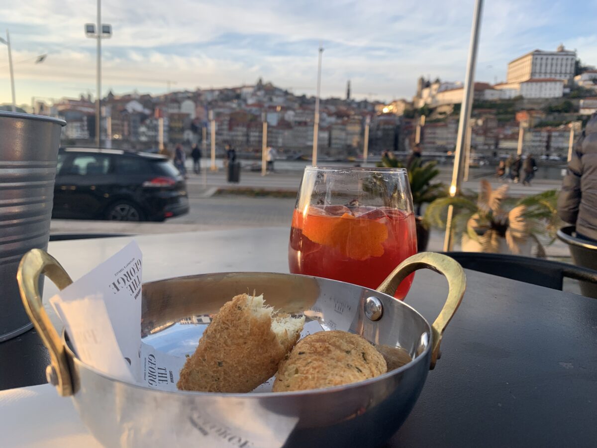 A bowl of fried fish cakes sits on a table in front of a glass of a red port cocktail with the blurry image of Porto's river and city in the distance