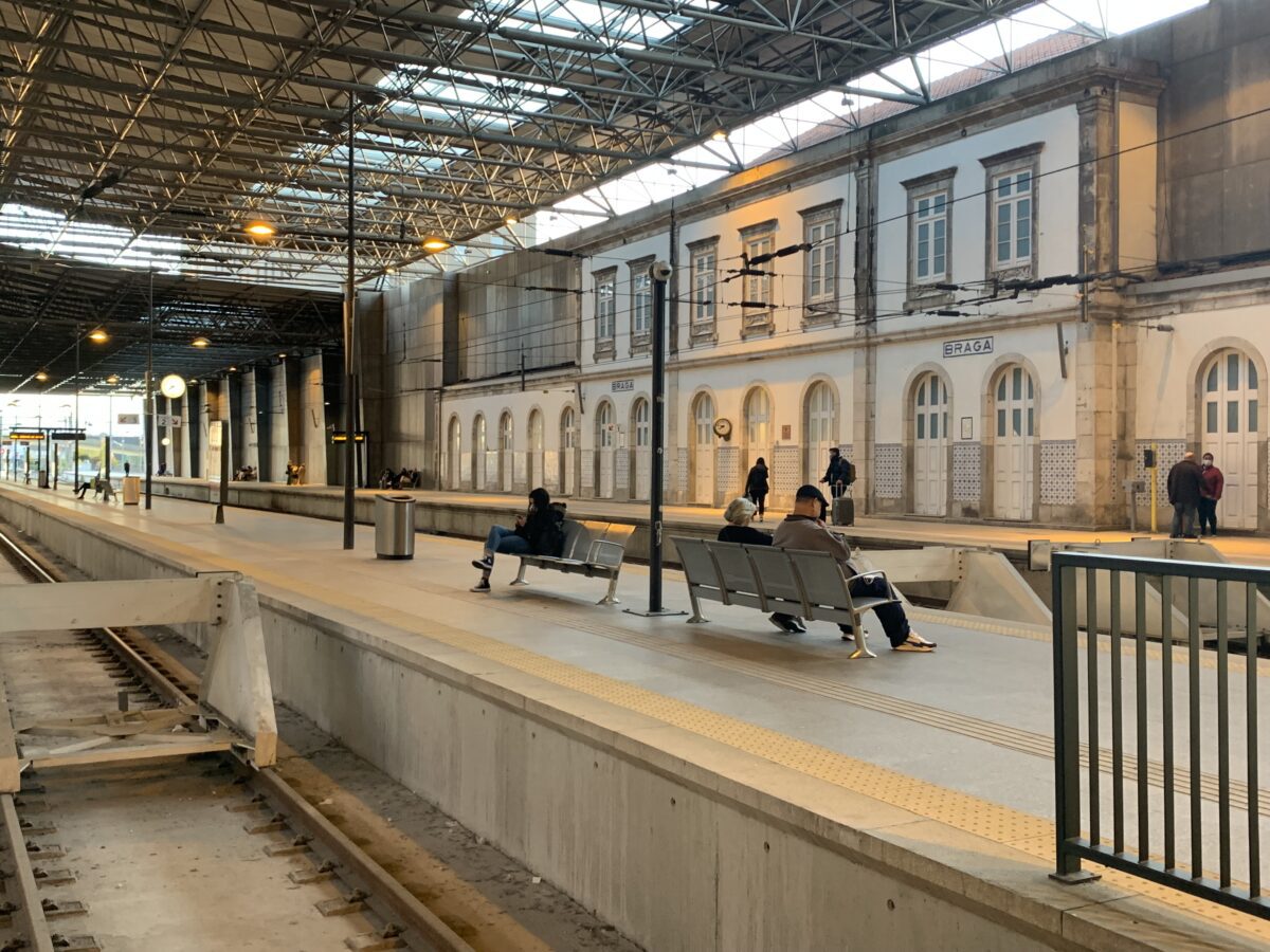 A train station platform with houses on the far side and benches with people sitting on them next to the lowered track area