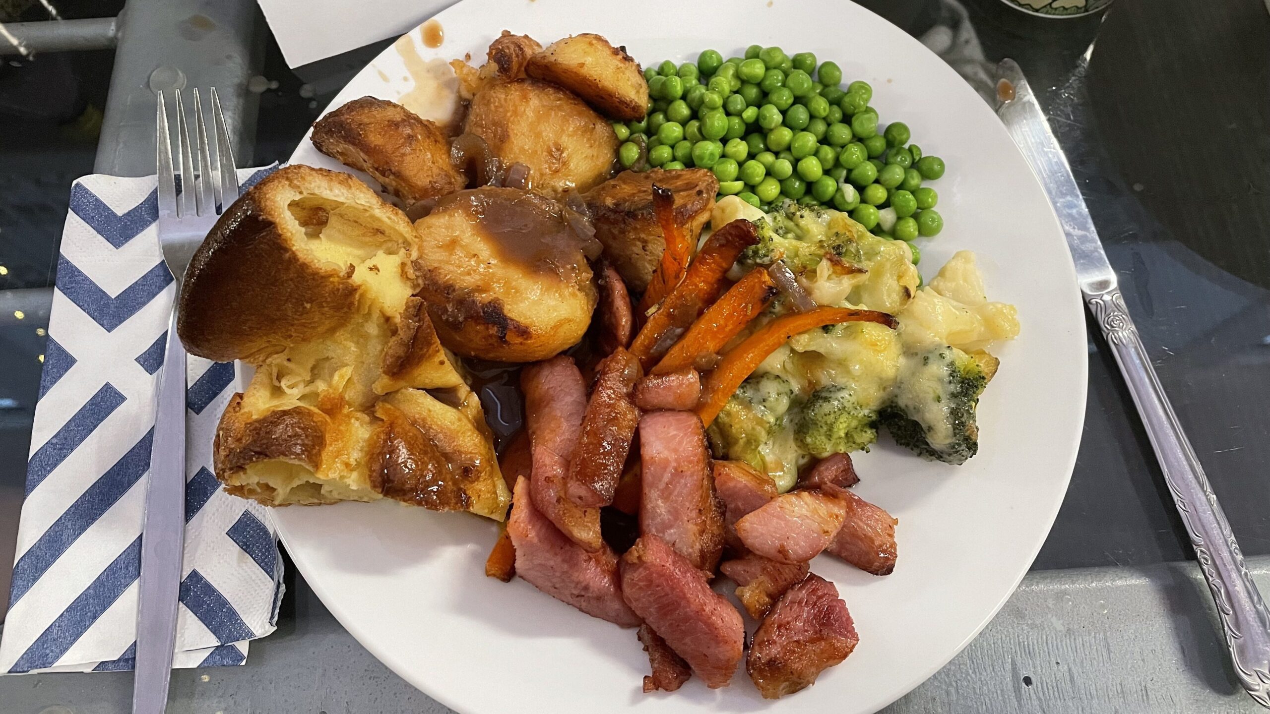 A plate of English roast with yorkshire pudding, peas, carrots, roasted potatoes, ham, and broccoli 