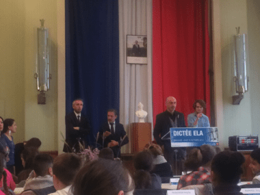 officials in suits stand behind a podium with large curtains making up the french flag behind them