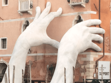 Two large statue hands rise from the canal and appear to hold up pink venetian buildings