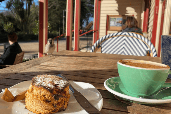 A green cup of chai latte and a pastry on a white plate sit on a wooden table with red and off-white wooden pillars and exterior paneled walls of a house porch with a small brown bird perched on the table's edge