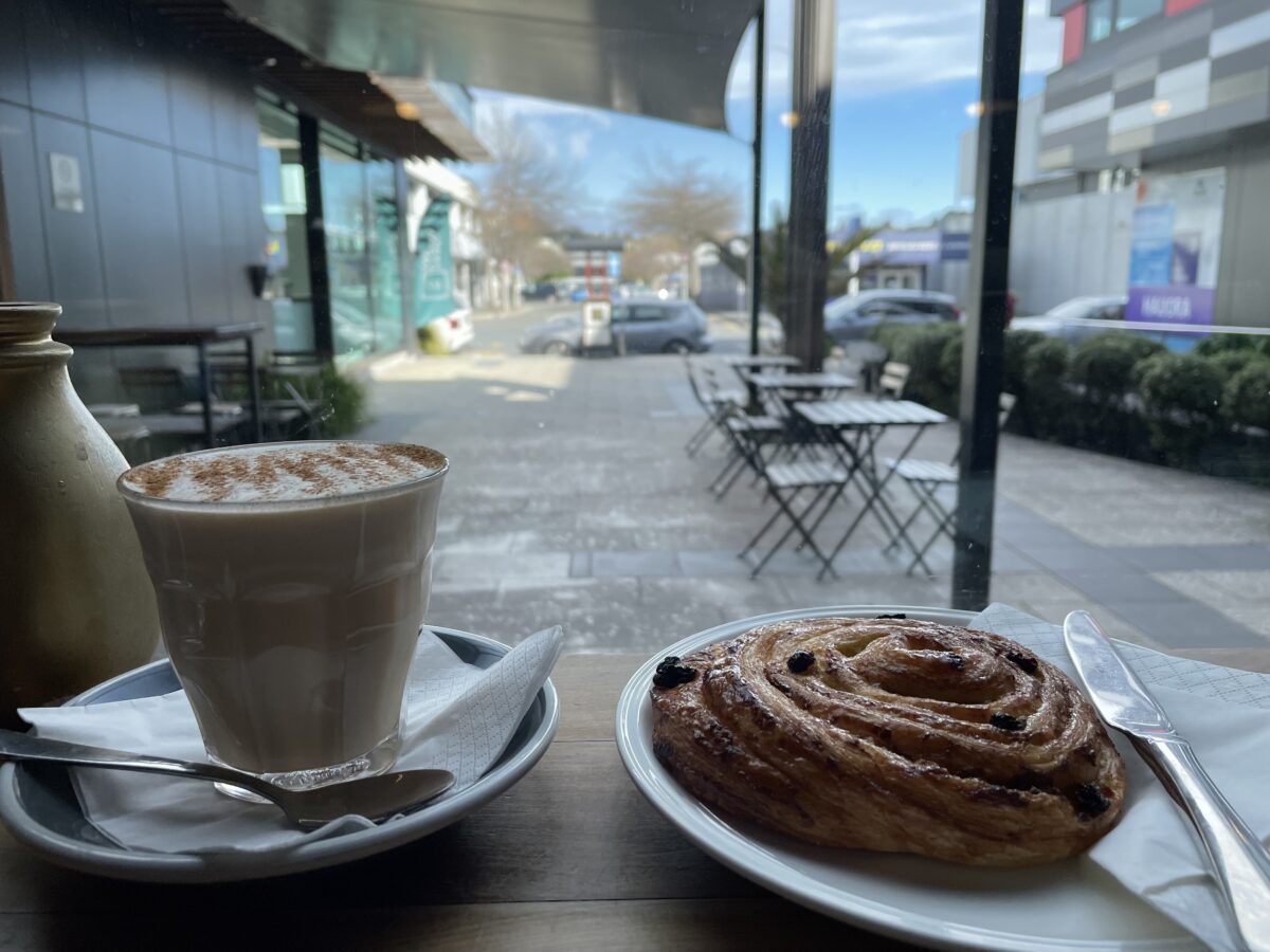 A chai latte in a clear cup and a pain au raison sit in front of a window looking over a cement square