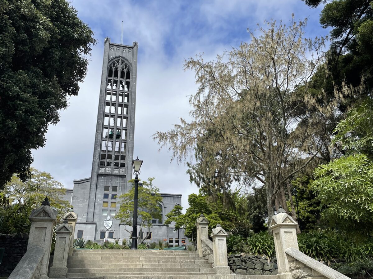 A tall concrete square tower of a cathedral with bells inside with trees on either side