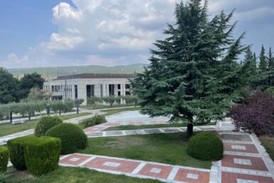 A campus lawn in Greece with red and white square tiles on the walkways between grass with cypress and olive trees. A stone library sits on the other side.