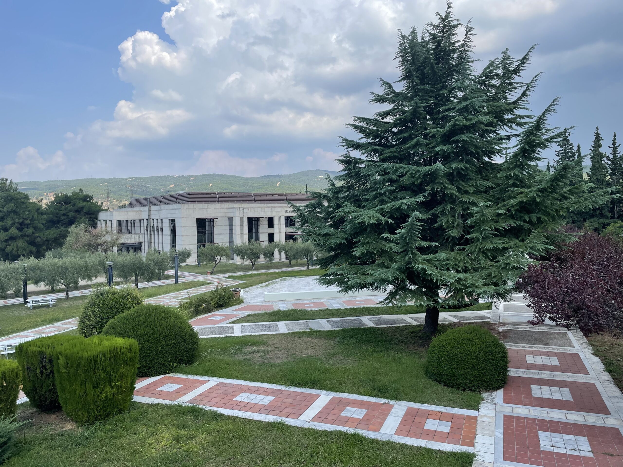 A campus lawn in Greece with red and white square tiles on the walkways between grass with cypress and olive trees. A stone library sits on the other side.
