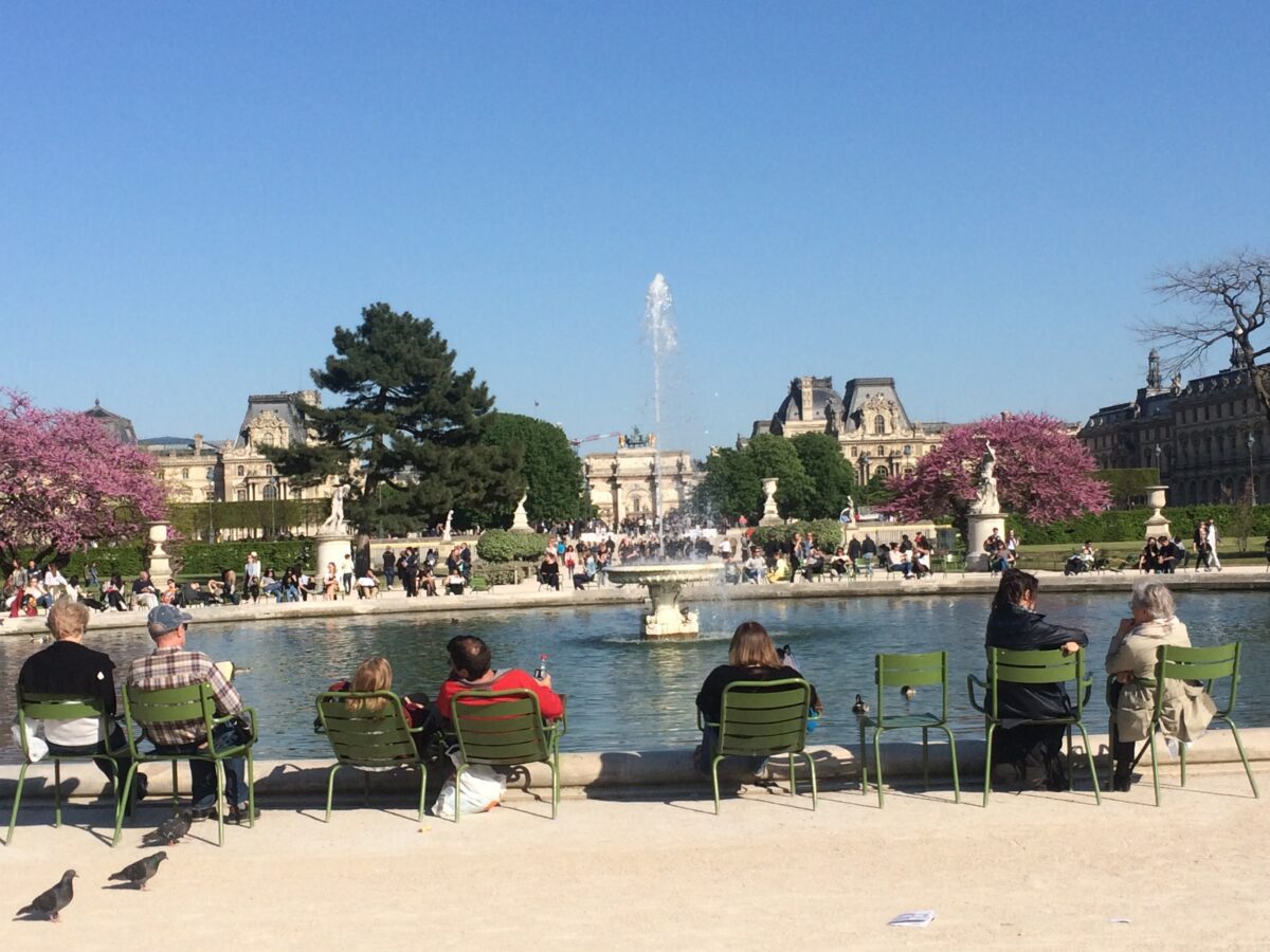 People sitting in garden chairs facing away from the camera and towards a large fountain with the louvre in the background