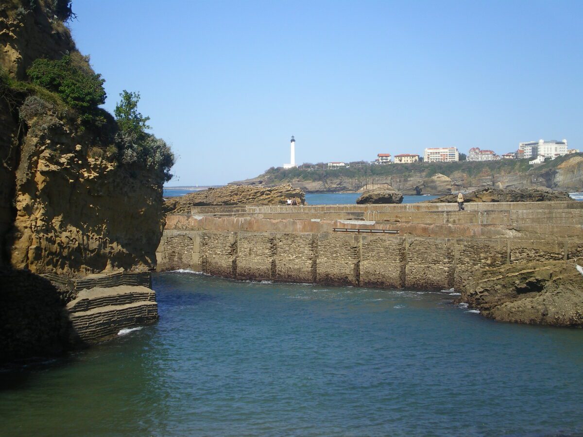 A harbor with a rock outcrop on the left, a stone sea wall down the middle, and a rocky shore in the background with a lighthouse