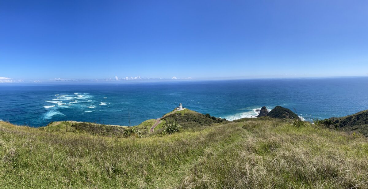A panorama showing whitecaps in the ocean in the background with a silhouette of rolling green hills and rocks in foreground with a lighthouse in the middle