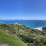 A walking path on the left, the outline of green hills stretch to the right with a view of the Cape Reinga lighthouse