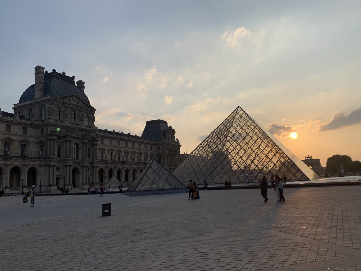 An arm of the louvre with the silhouette of the glass pyramid in front with the orange sunset in the back