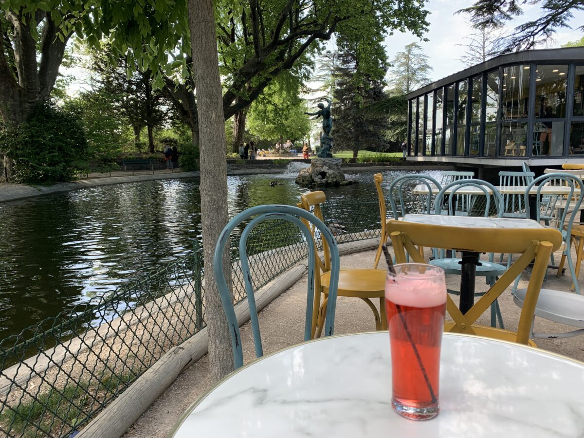 A red drink sits on a cafe table outside with a pond surrounded by trees