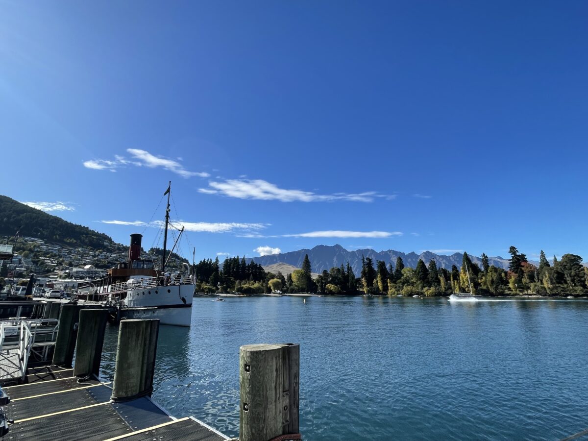 Lakefront photo from a wharf with a large white and red steamboat on the left and the Queenstown lake front on the other side of the lake with the Remarkables mountain range in the background