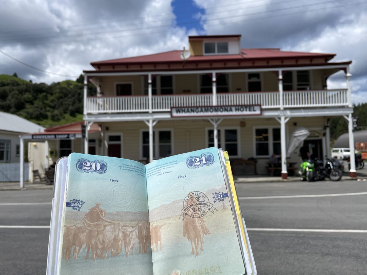 A US passport is open to a page with a stamp for Whangamomona in front of the Whangamomona Hotel on the Forgotten World Highway