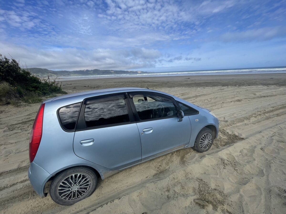 A light blue hatchback car is stuck in the sand facing towards the beach.