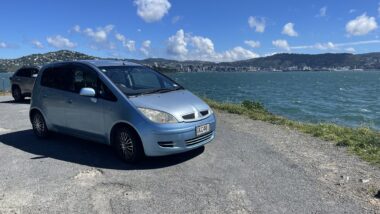 A light blue hatchback car parked on the side of a gravel road next to the sea.