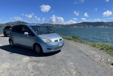 A light blue hatchback car parked on the side of a gravel road next to the sea.