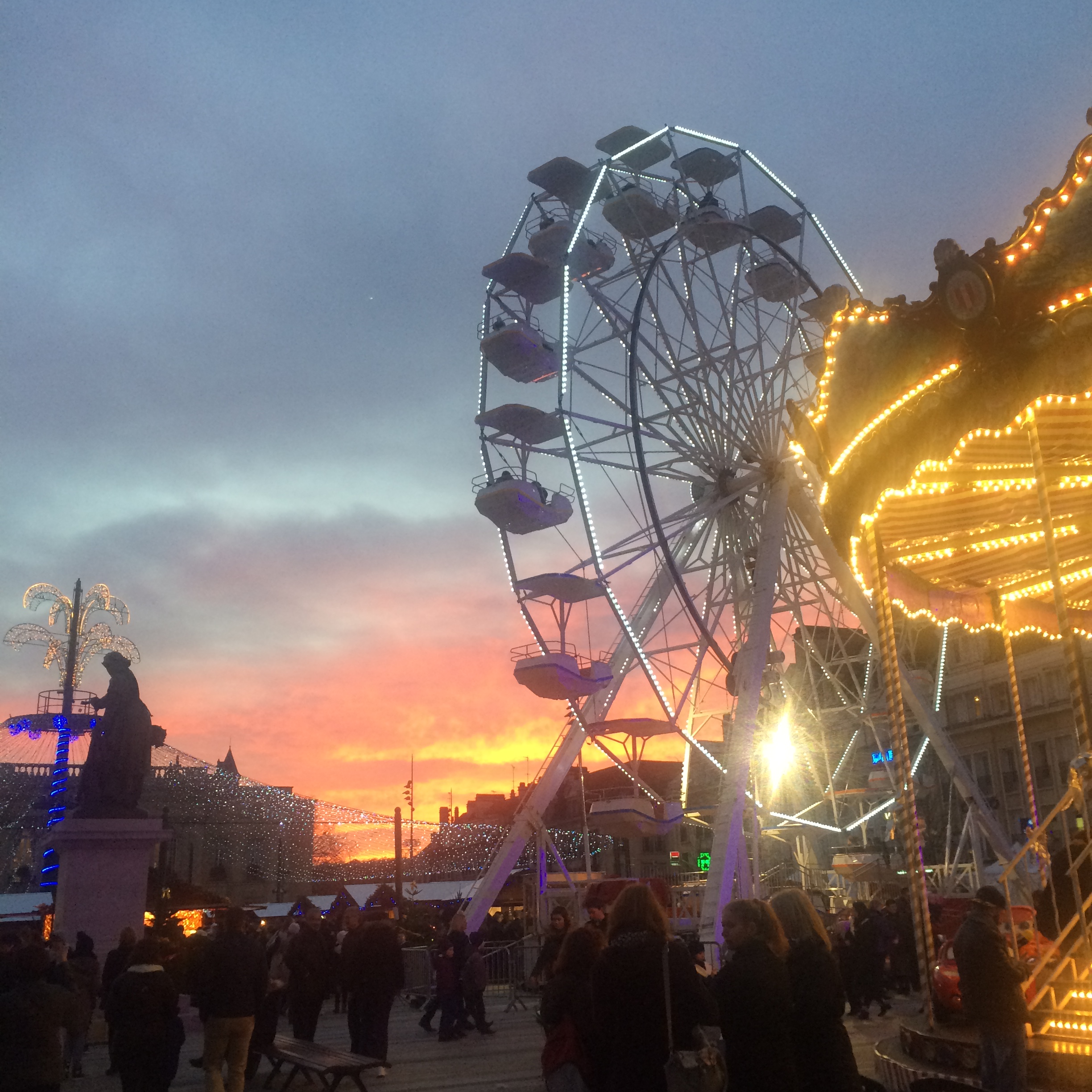 During sunset, a ferris wheel, carousel, and winter market is lit up in the city square