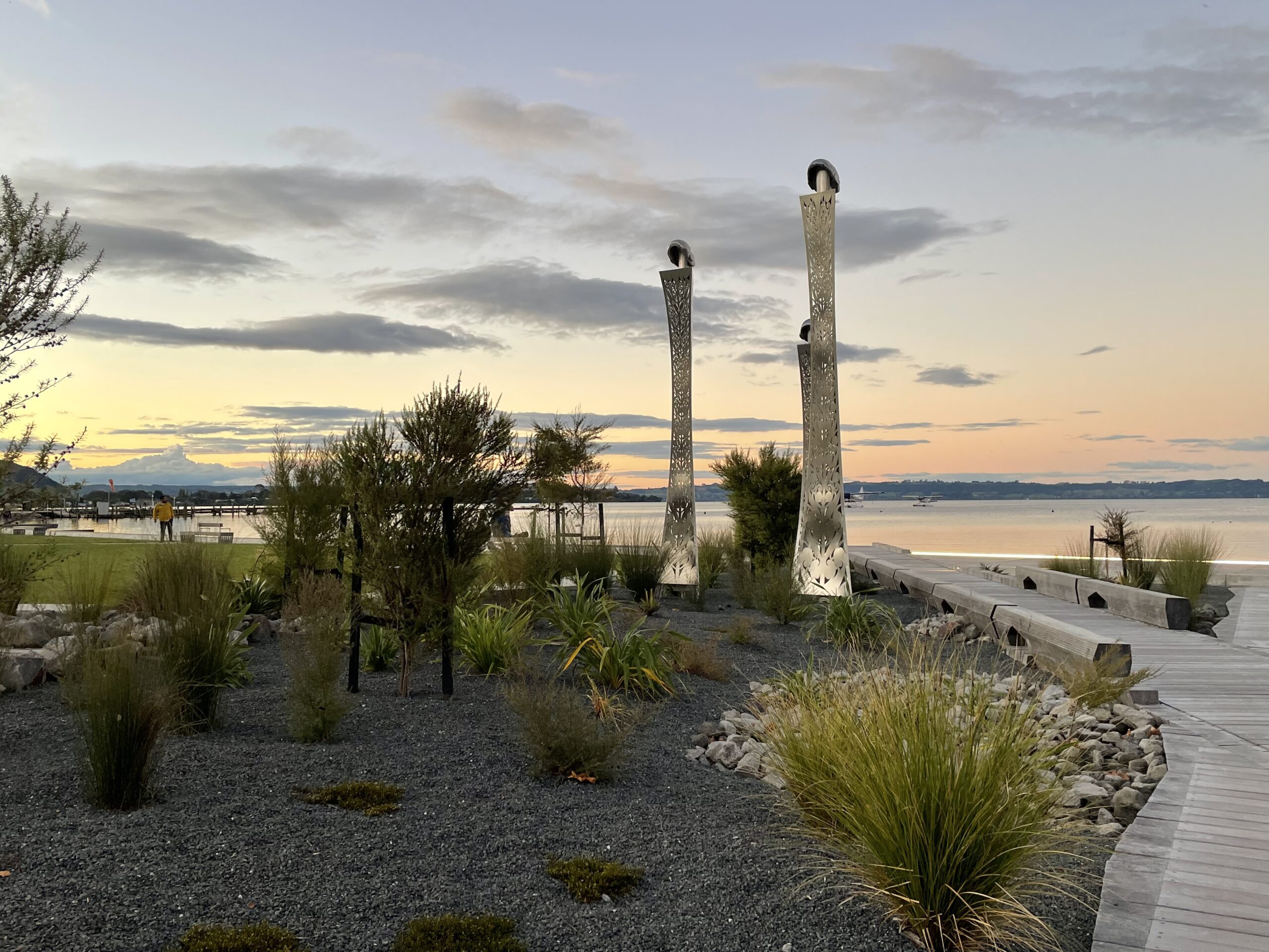 Wooden walkway leading to a lake at sunset with sparse bushes and two tall totems