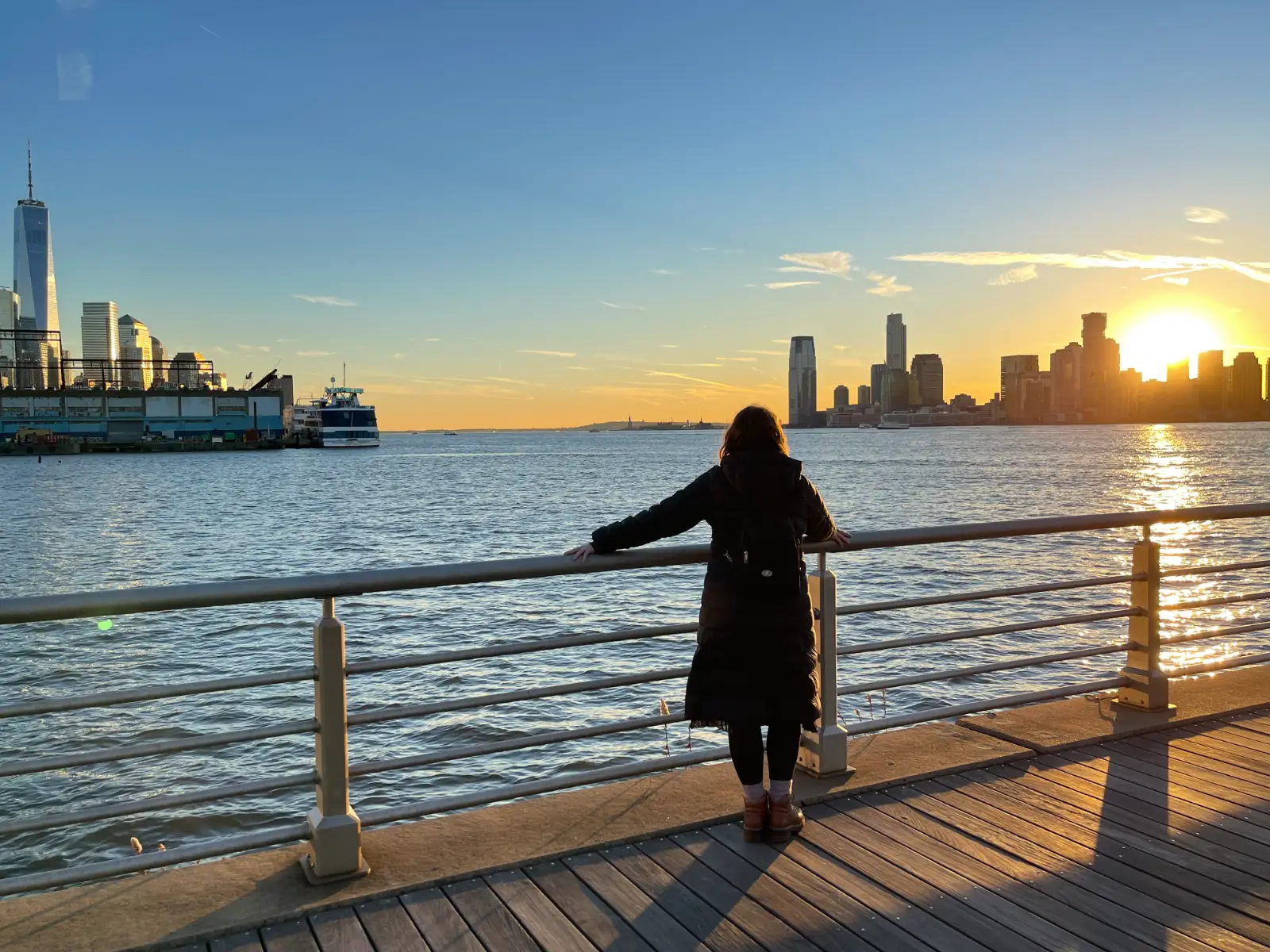 Silhouette of a woman standing at a guardrail in front of a river with the edge of Manhatten and Jersey City skylines in the distance during sunset